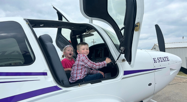 Two children smile while sitting inside of a K-State aircraft.